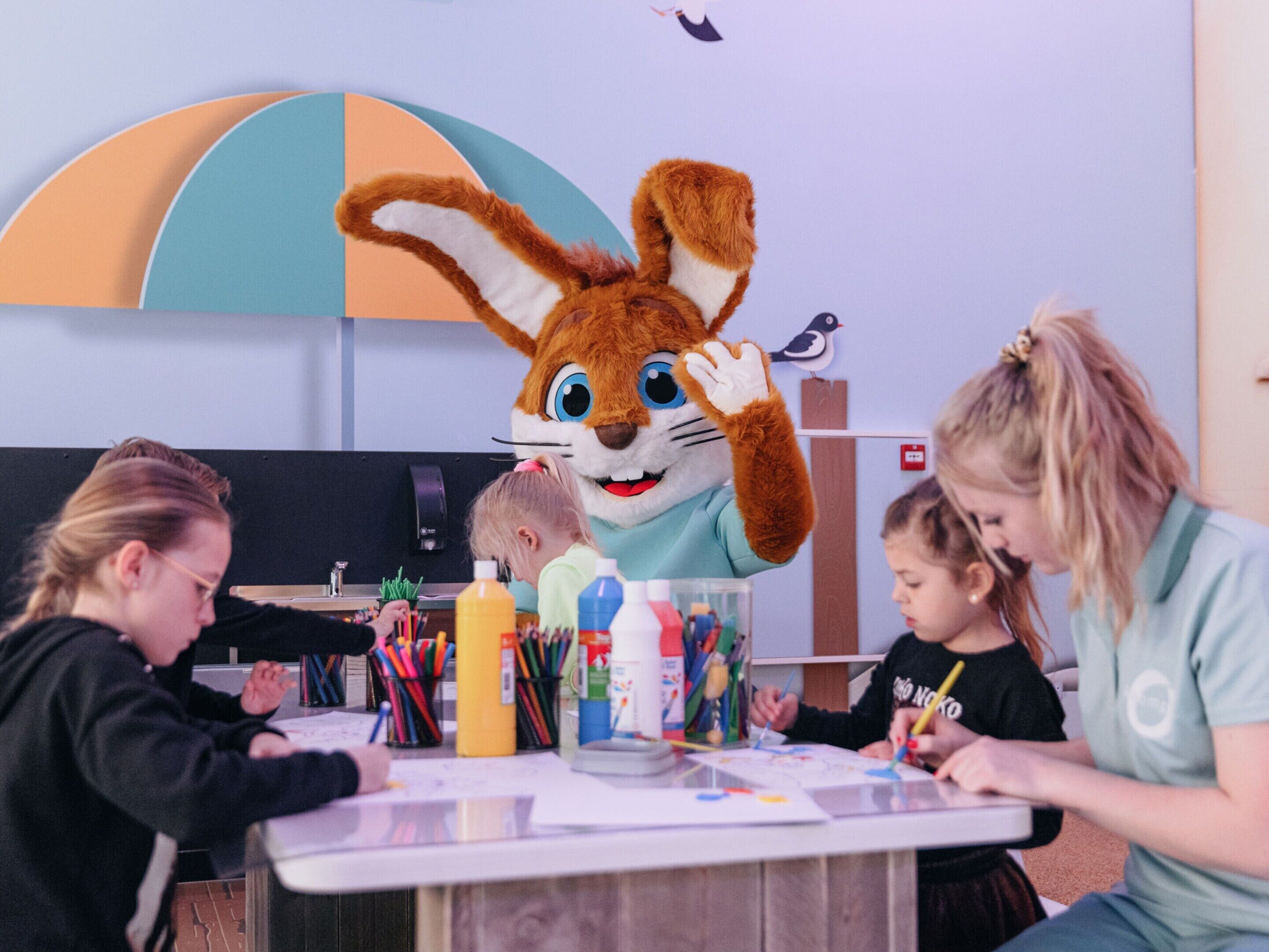 A classroom with a mascot, children and adults engaged in drawing activities.