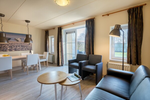 Una sala de estar moderna con un sofá, sillas, una mesa de centro y un comedor con mesa y sillas. Hay ventanas grandes con cortinas, una lámpara de techo y un cuadro en la pared.