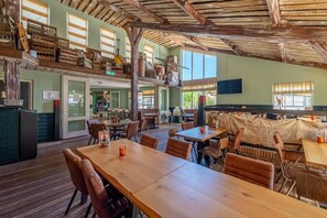 Un vaste espace dédié à la salle à manger, avec des tables et des chaises en bois, un plafond haut avec des poutres apparentes et une vue sur l’espace extérieur aménagé pour les repas.