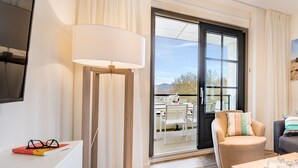 Una habitación de hotel moderna con un gran ventanal corredero que da a un espacio al aire libre, una lámpara de pie, un sofá con cojines y una mesa con una bandeja de vasos.