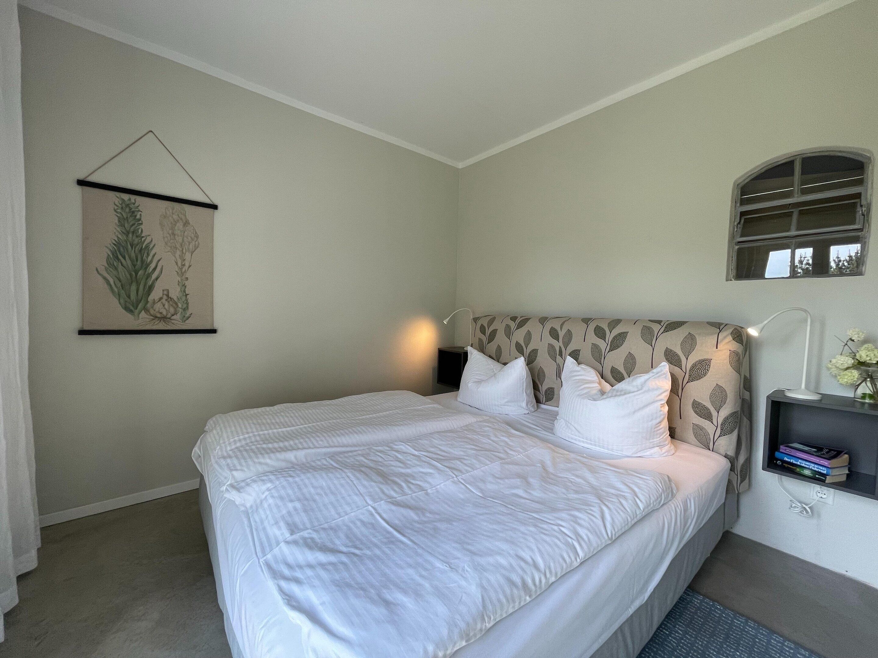 A neatly made bed with white bedding, a headboard with a leaf pattern, a bedside table with a lamp, and a framed botanical print on the wall.