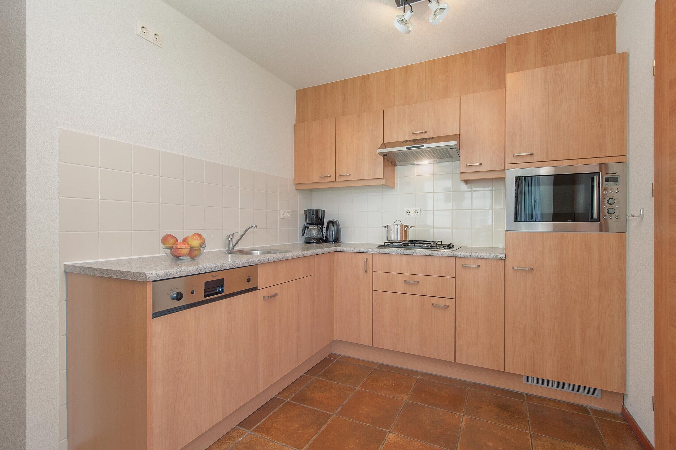 Una cocina con armarios de madera, microondas, hervidor de agua y un bol de fruta en la encimera.