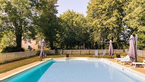 A rectangular swimming pool with clear blue water, surrounded by a wooden fence, trees, and lounge chairs with umbrellas. - Cottage in Chaleix Near Forest & Lake (Chalais)