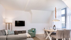 Een moderne woonkamer met een grijze bank, een groene fauteuil, een eettafel met stoelen, een flatscreen televisie en een lamp.