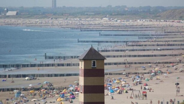 Een strand met een vuurtoren op de voorgrond, een pier die zich uitstrekt over het water en een verafgelegen stadshemel.