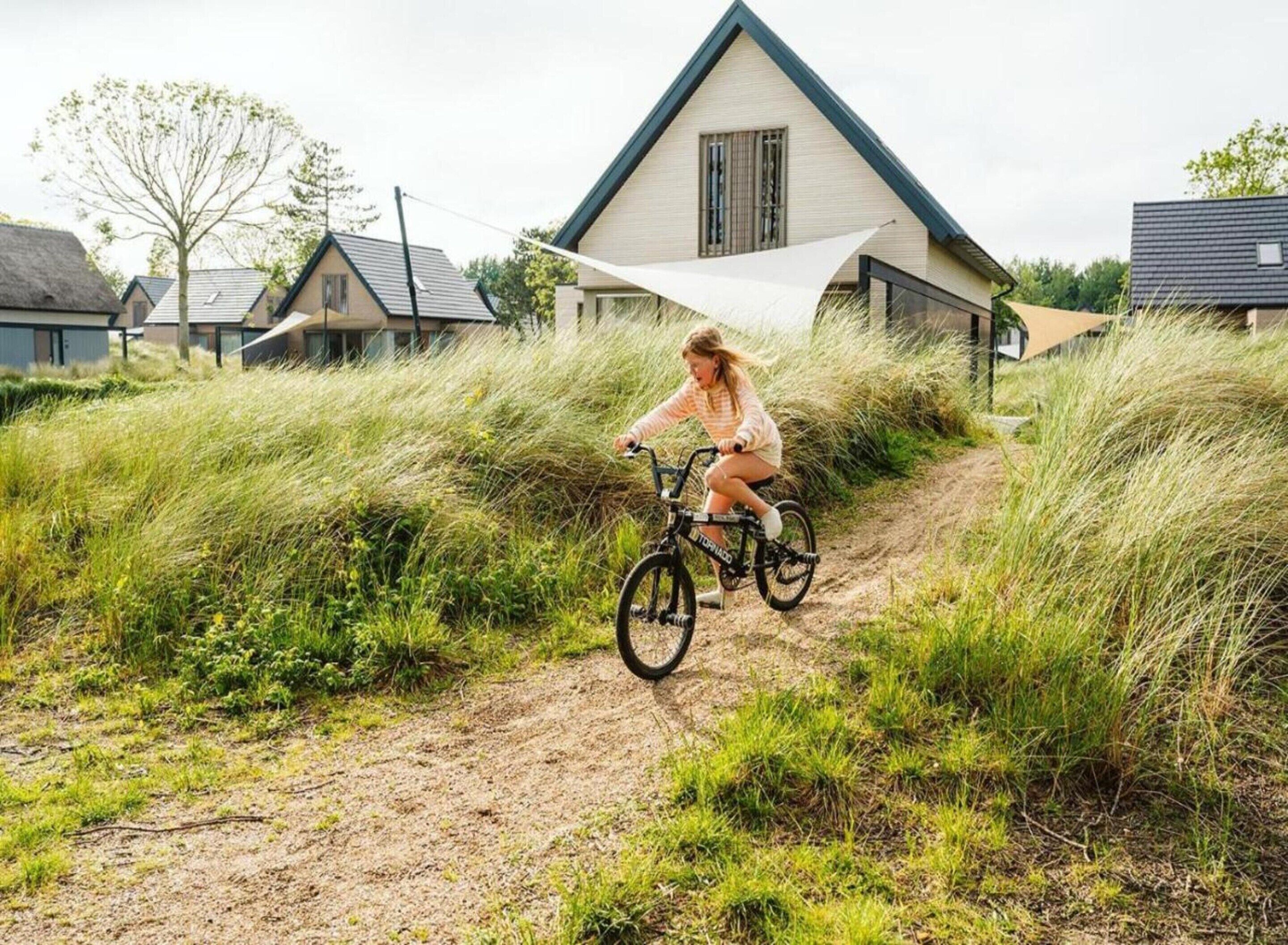 A person riding a bicycle on a dirt path surrounded by tall grass and houses with modern architecture.