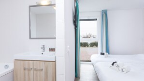 Une chambre d’hôtel avec un meuble en bois, un lavabo blanc, un miroir, un lit avec une literie blanche et une vue par la fenêtre.