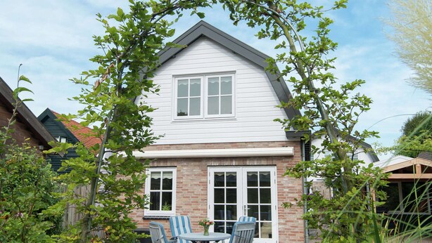A two-story house with a brick foundation and white siding, featuring a patio with a table and chairs, surrounded by greenery. - Comfortable Flat in Bergen (Bergen)