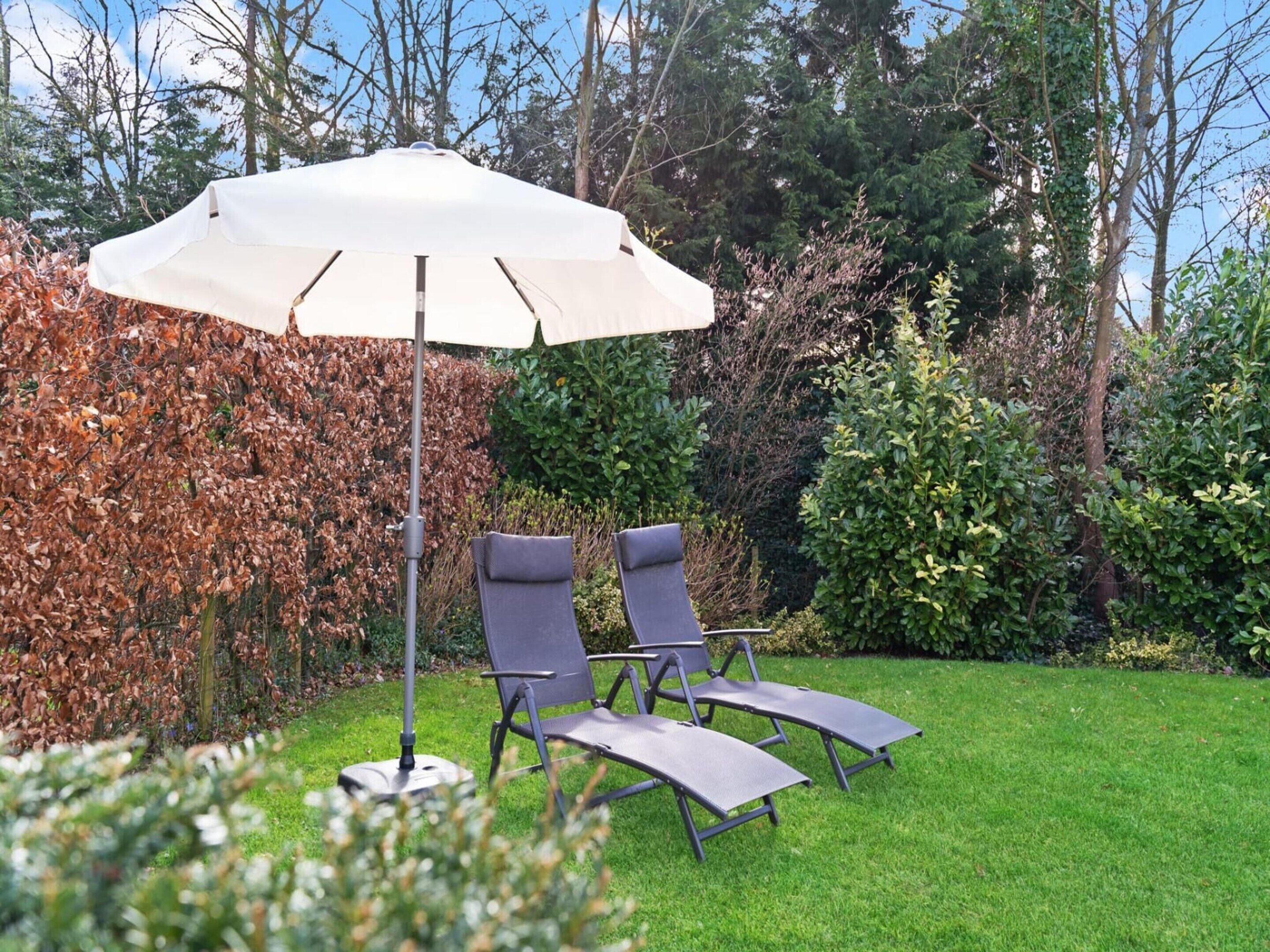 A garden with a white umbrella, two lounge chairs, and a hedge.