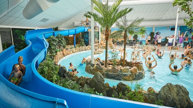 An indoor water park with a blue water slide, a central pool surrounded by rocks and palm trees, and people swimming and playing. - Villa With Sauna & Bubble Bath in Limburg (Roggel)