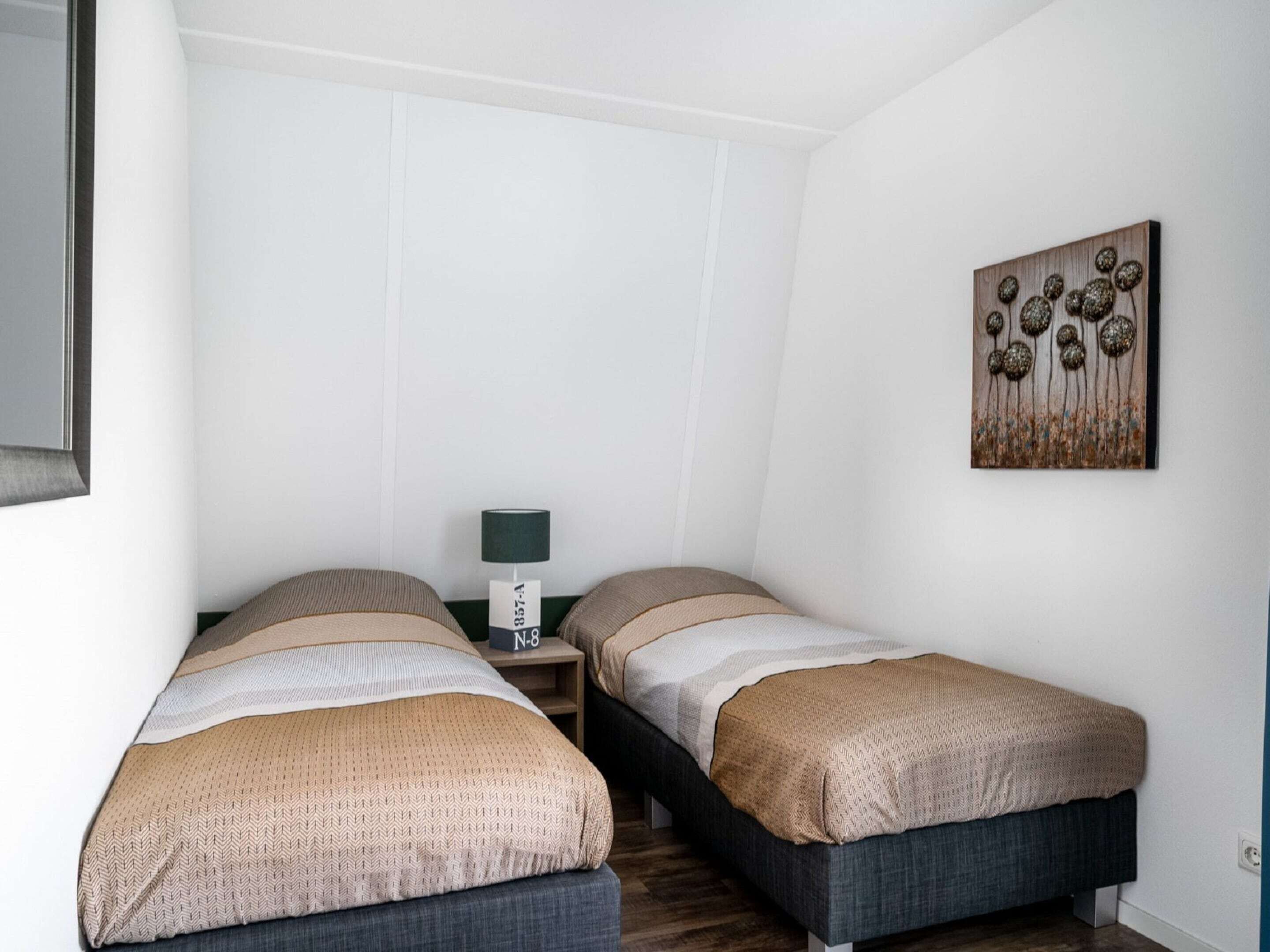 Een kleine, moderne slaapkamer met twee eenpersoonsbedden, een nachtkastje met een lamp en een kunstwerk aan de muur.