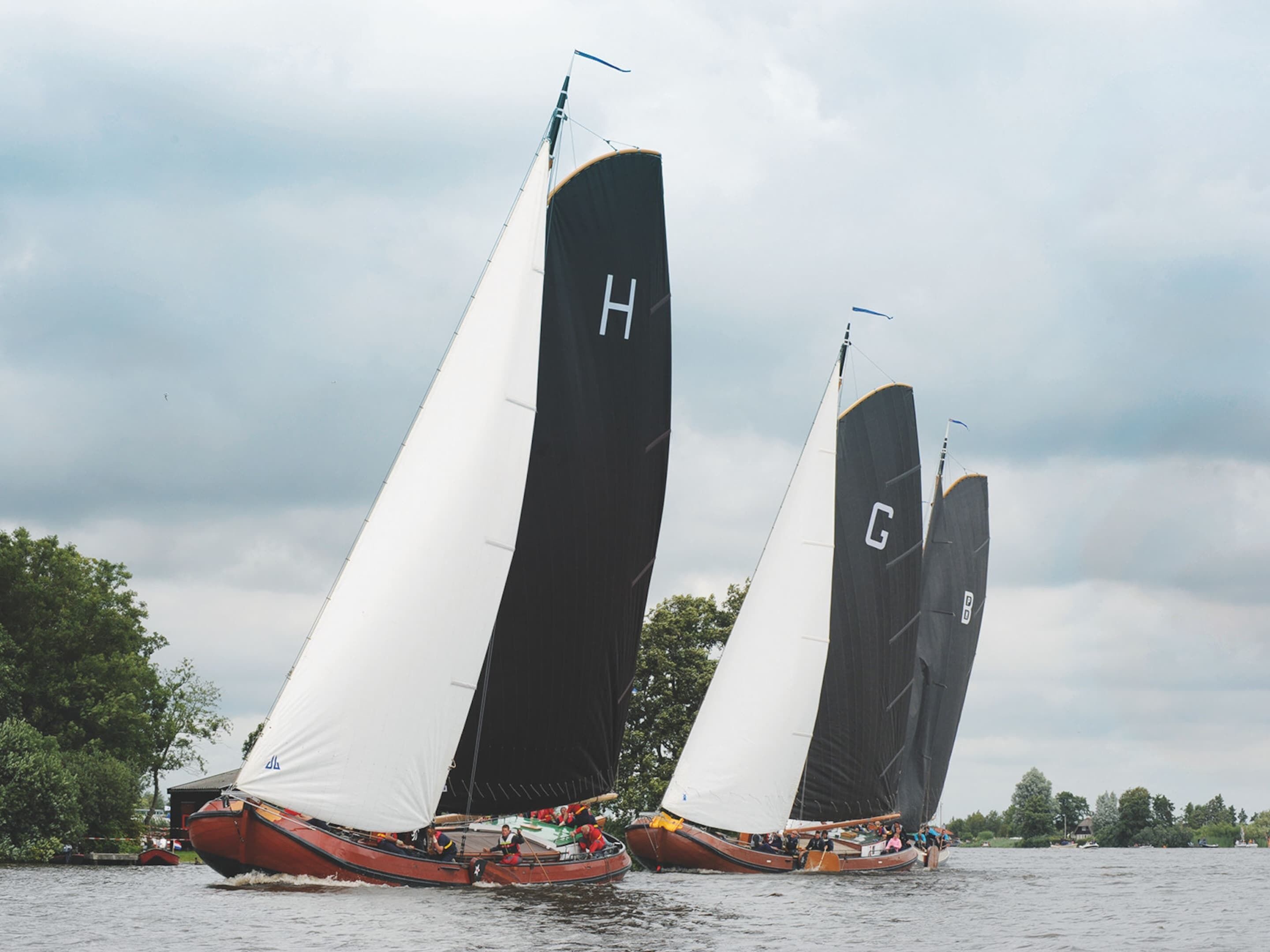 Two sailboats with black sails marked with letters H and G on a body of water.