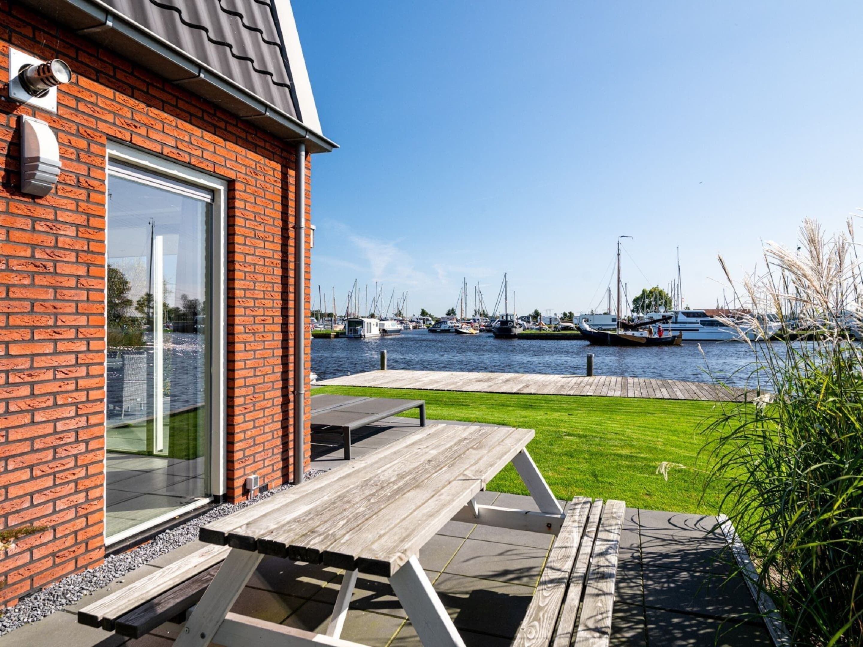 A patio with a table and chairs, a brick building with a glass door, and a marina with boats in the background.