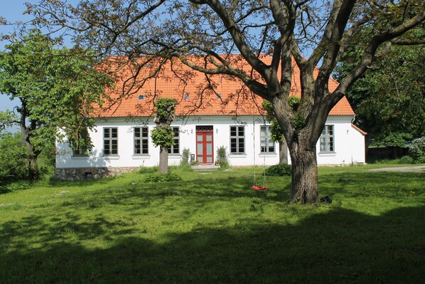 A white building with a red door and a red roof, surrounded by trees and grass. - Ferienwohnung in Steffenshagen mit Garten (Steffenshagen)