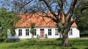 A white building with a red door and a red roof, surrounded by green grass and trees.