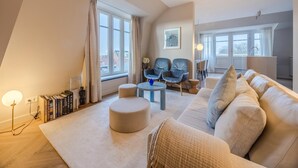 Ein modernes Wohnzimmer mit großem Fenster, einer Couch, zwei Sesseln, einem runden Hocker und einem Bücherregal.