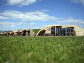Una hilera de edificios modernos con techos de madera y grandes ventanales, frente a un campo verde y un cielo despejado.