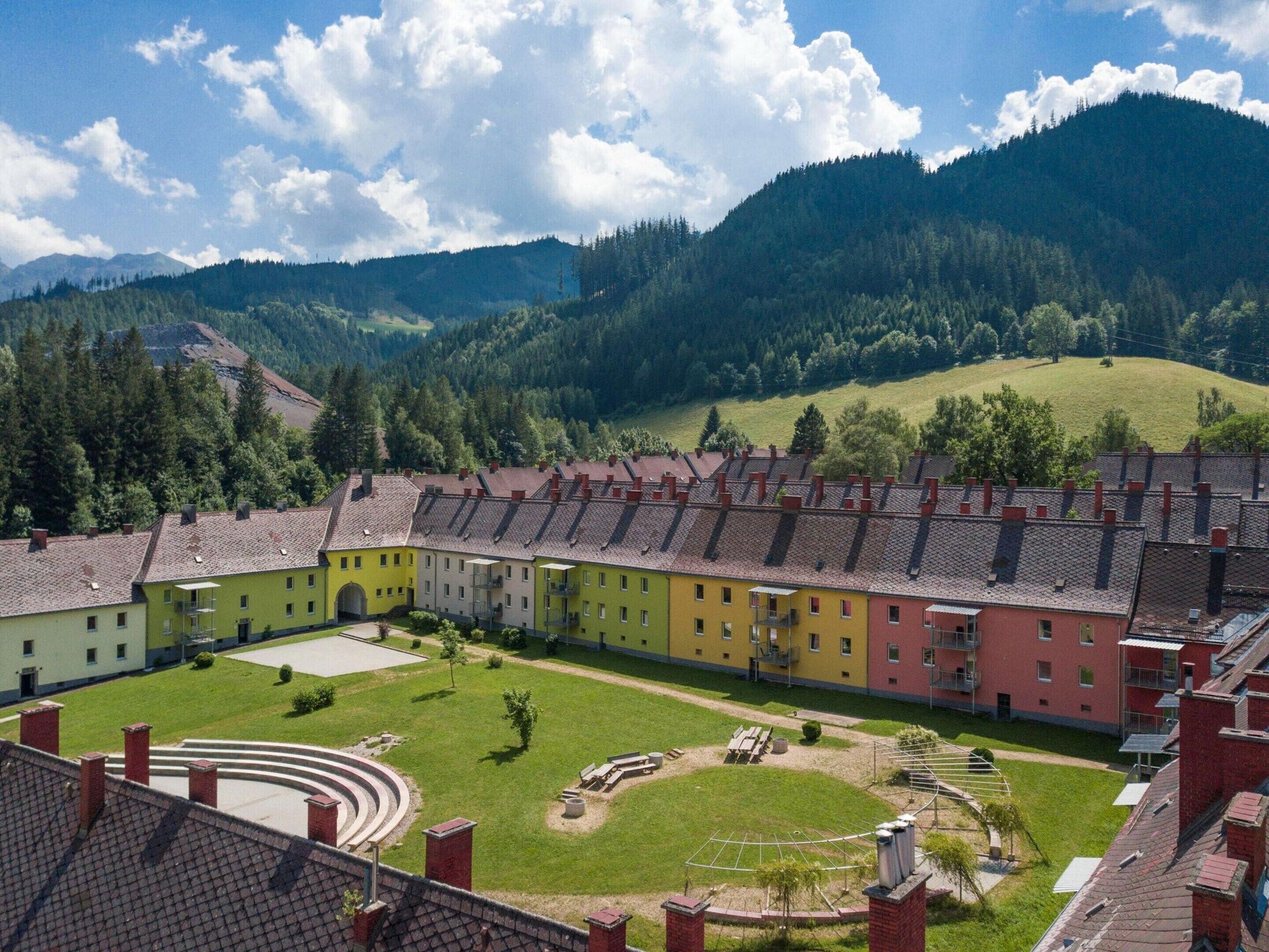A row of multi-story buildings with a courtyard in the center, surrounded by greenery and mountains.