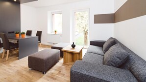 A modern living room with a grey sofa, a wooden coffee table, and a dining area with a table and chairs. There is a window providing natural light and a view of greenery outside.