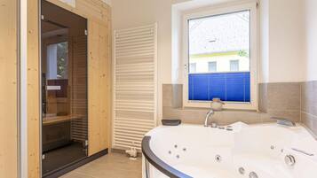 Un baño moderno con una bañera grande, una ventana con persianas y una puerta de madera que da a una zona de ducha.