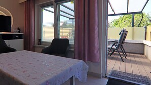 A room with a table covered in a floral tablecloth, a television on a stand, and a window with curtains. - Bungalow Near Baltic Sea (Neubukow)