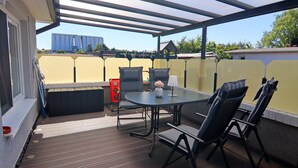 A covered patio with a table and chairs, a small birdhouse, and a view of a large industrial building in the background. - Bungalow Near Baltic Sea (Neubukow)