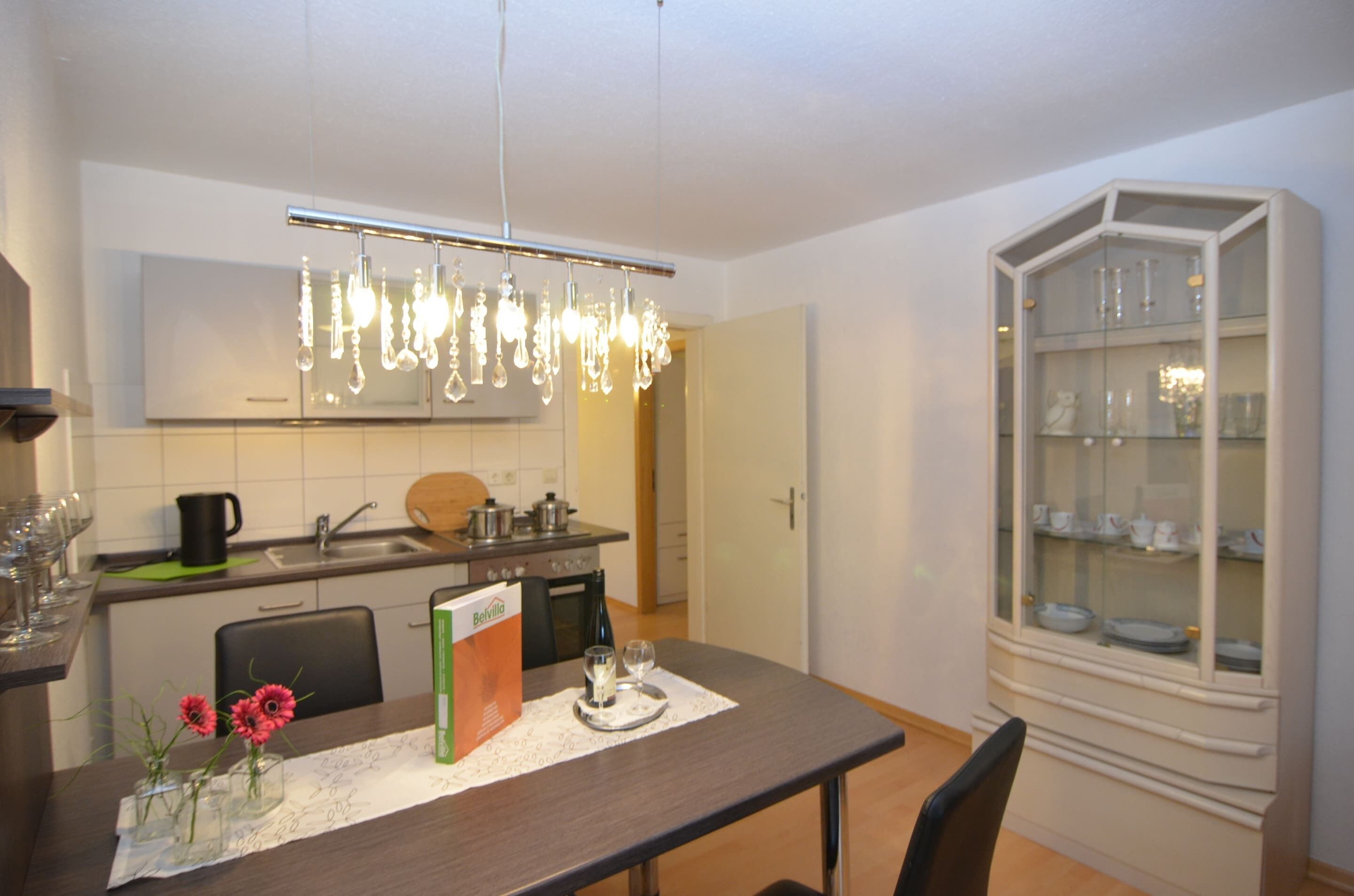 Une cuisine moderne avec un coin repas, agrémentée d’un lustre, d’un meuble avec des portes en verre et d’une table dressée pour deux personnes.