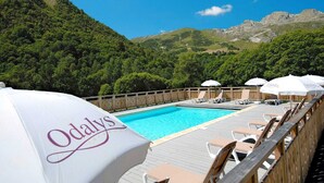Ein Swimmingpool mit Liegestühlen und Sonnenschirmen, umgeben von einer Holzterrasse und üppiger Bepflanzung.