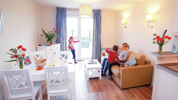 Ein Wohnzimmer mit Sofa, einem Esstisch mit Stühlen, einer Küchenzeile und Blick durch ein Fenster.