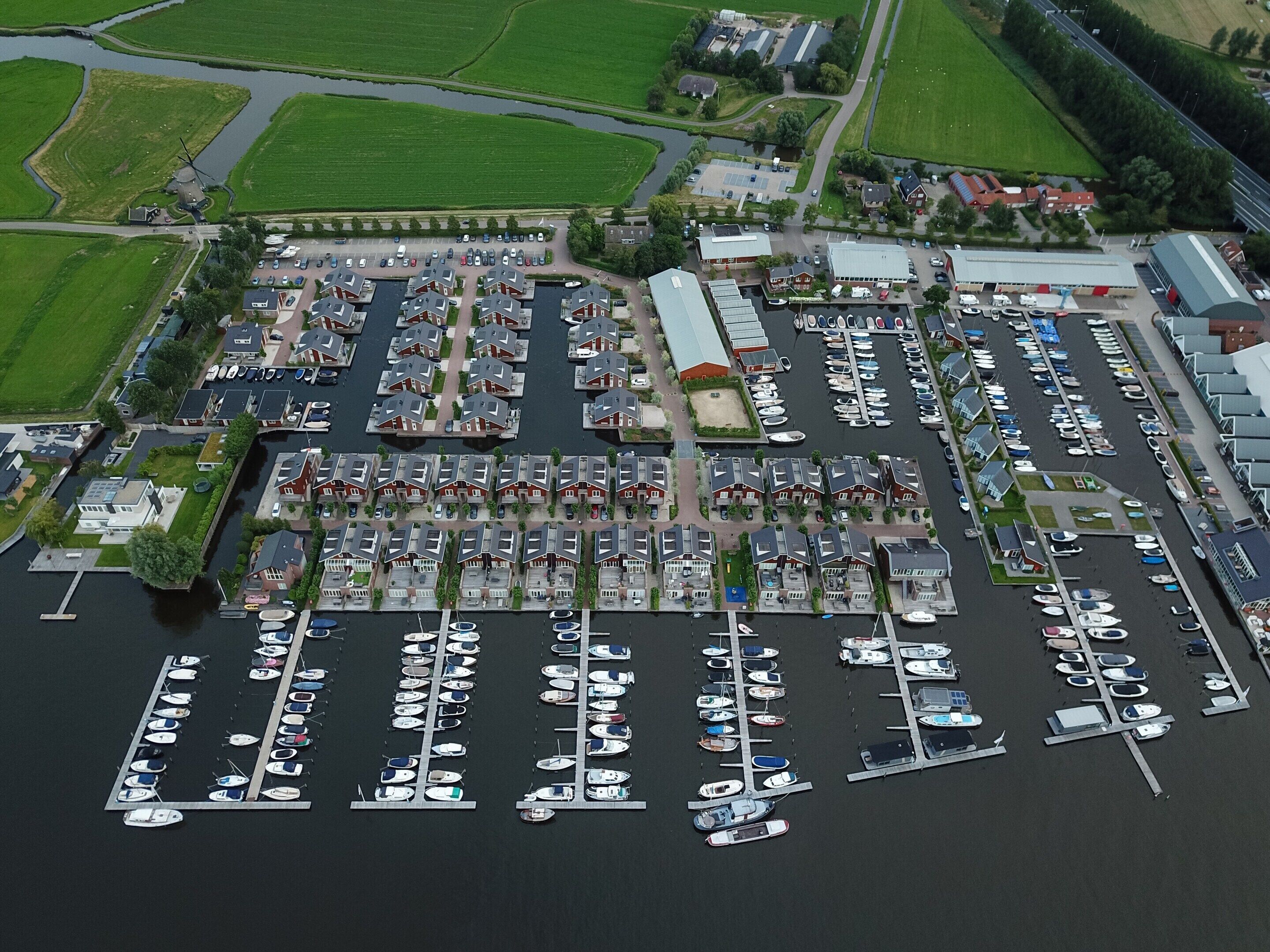 Une vue aérienne d’une marina où de nombreux bateaux sont amarrés selon un plan bien défini, entourée de bâtiments résidentiels et d’un vaste complexe industriel.