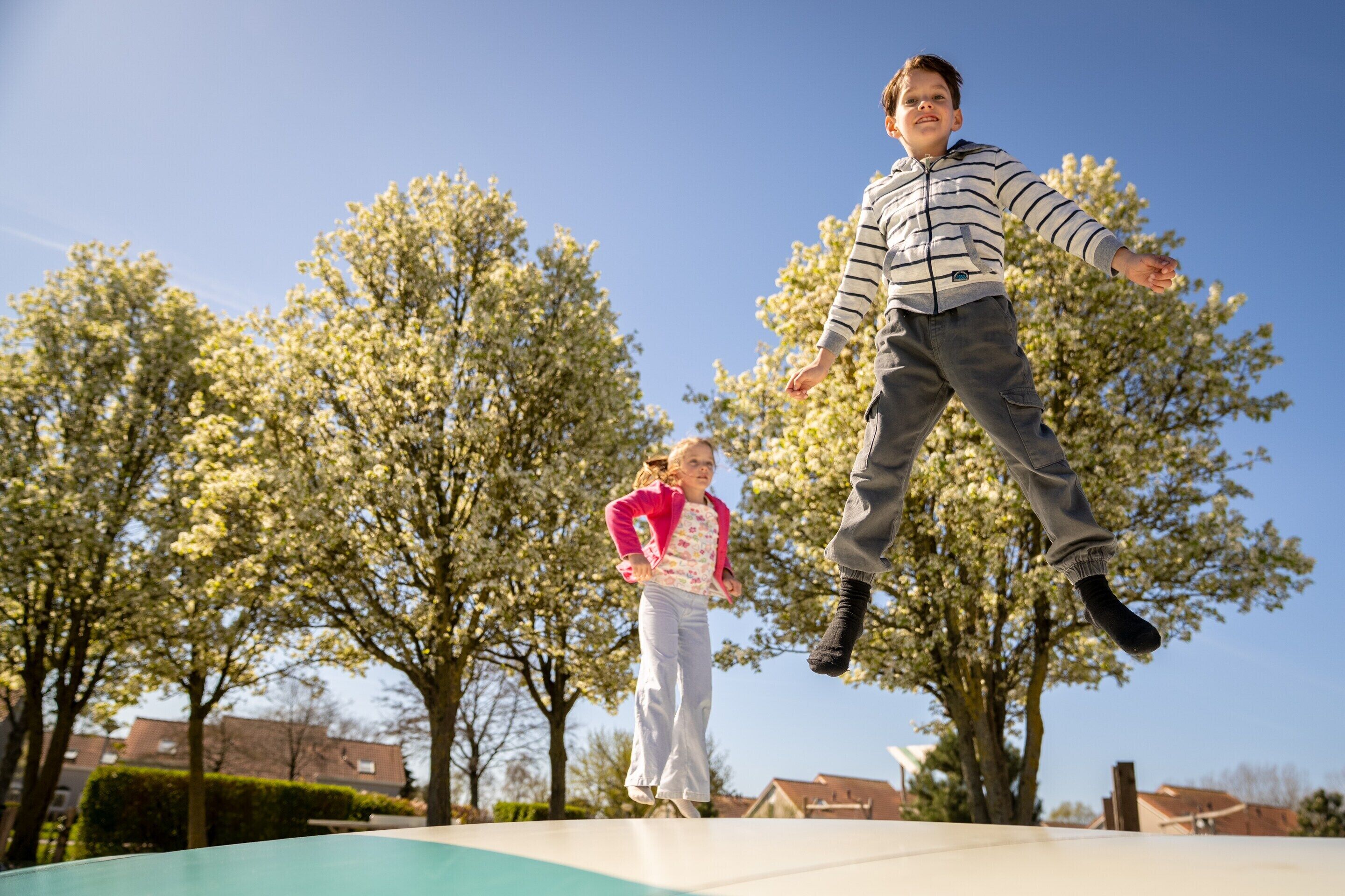 Una persona saltando en un trampolín al aire libre, con árboles y casas al fondo.