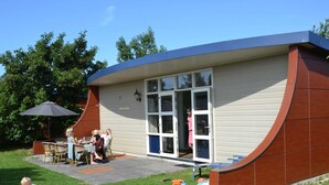 A small, single-story building with a curved roof, a patio with outdoor seating, and a large umbrella. - Bungalow in Dunes With Nautical Charm ('s-Gravenzande)