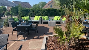 Un espace extérieur aménagé en terrasse, avec des coins salon comprenant un canapé et des fauteuils, ainsi qu’une table à manger.