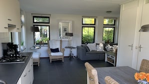 Une cuisine moderne équipée d’une cuisinière à gaz, un coin repas avec des chaises en osier, un canapé gris et de grandes fenêtres offrant une vue sur la verdure.