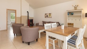 A spacious living room with a fireplace, a dining area with a wooden table and chairs, a sofa, and a bookshelf. - Bungalow by the Sea With Sauna in Germany (Sierksdorf)