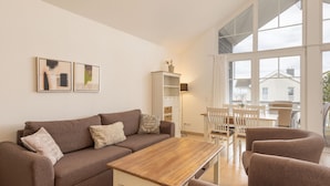 A modern living room with a brown sofa, a wooden coffee table, and white chairs. There are framed pictures on the wall and a cabinet with shelves. - Bungalow by the Sea With Sauna in Germany (Sierksdorf)