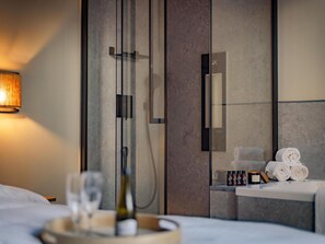 Una habitación de hotel moderna con cama, una mesita de noche con un vaso y una botella, un perchero con toallas y una zona de ducha con puerta de cristal.