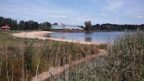 Une zone au bord d’un lac, avec un b âtiment doté d’un toit à rayures, entouré d’arbres et bordé d’une plage de sable.