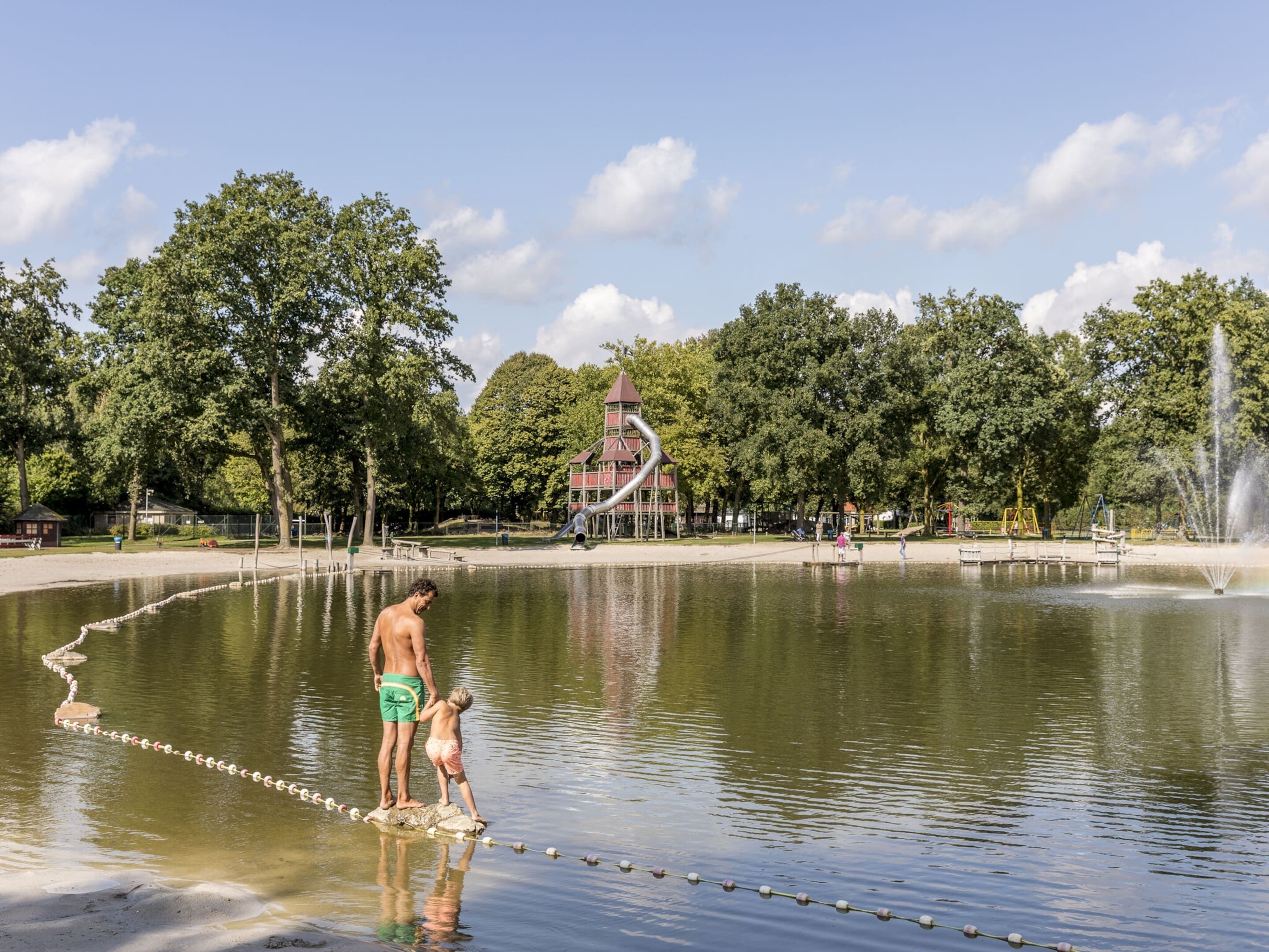 Un homme et un enfant se tiennent sur un pont de corde qui enjambe une étendue d’eau dans un parc. On aperçoit en arrière-plan des équipements de jeux pour enfants et une structure en forme de tour.