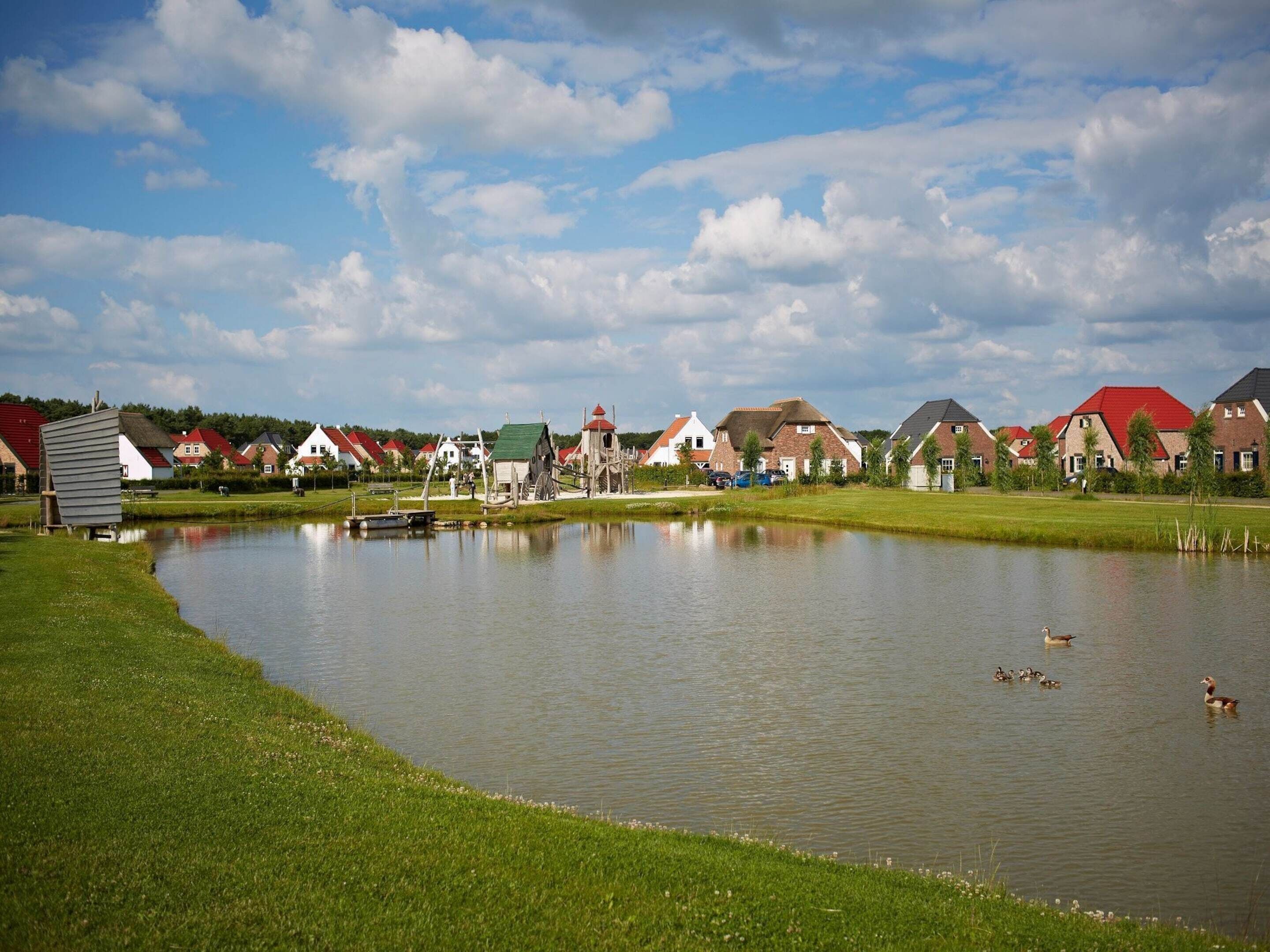 A serene lakeside setting with traditional houses, a small boat, and ducks swimming.