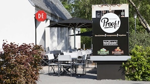 Un área de estar al aire libre con mesas y sillas, un letrero que dice 'De Boshoek Restaurant' y un letrero rojo con la inscripción 'DE'.