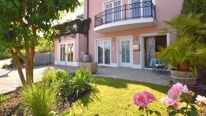 A pink building with a balcony, a garden with grass and plants, and a door with a window.