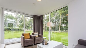 Un salon moderne doté d’une grande porte-fenêtre coulissante donnant sur un jardin. On y trouve deux fauteuils avec des coussins, une table basse blanche et un lampadaire.