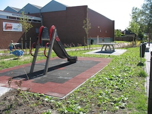 Ein Spielplatz im Freien mit Rutsche und Schaukeln, einem Picknicktisch sowie einem Gebäude mit Beschilderung im Hintergrund.
