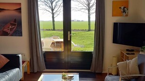 A living room with a sofa, a coffee table with a plant, a TV, and a view of a grassy field through a glass door. - Nice House with Dishwasher near Wadden Sea (Tzummarum)