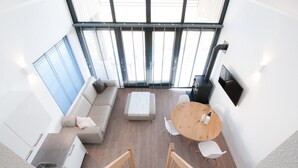 Un intérieur moderne comprenant un canapé, une table basse, des chaises, un coin cuisine et une grande fenêtre avec des stores.
