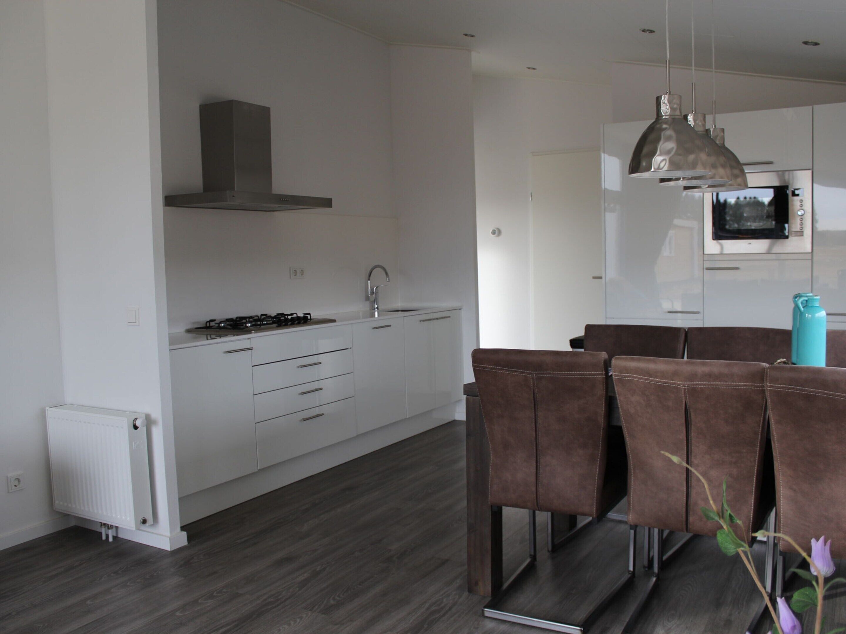 A modern kitchen with white cabinets, a gas stove, and a built-in oven. There is a dining area with brown chairs and a teal water bottle on the table.