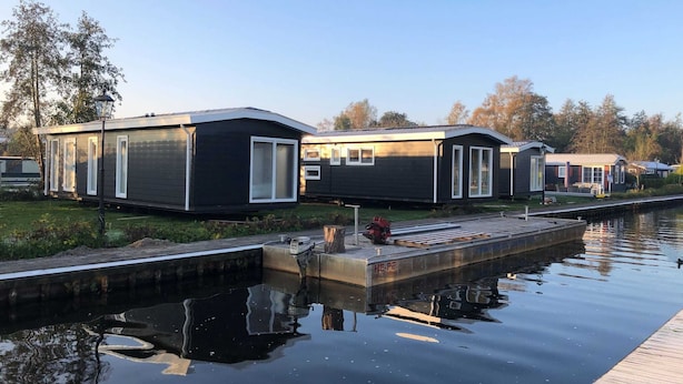Una hilera de casas móviles modernas de color oscuro con detalles blancos, ubicadas junto a un canal tranquilo.