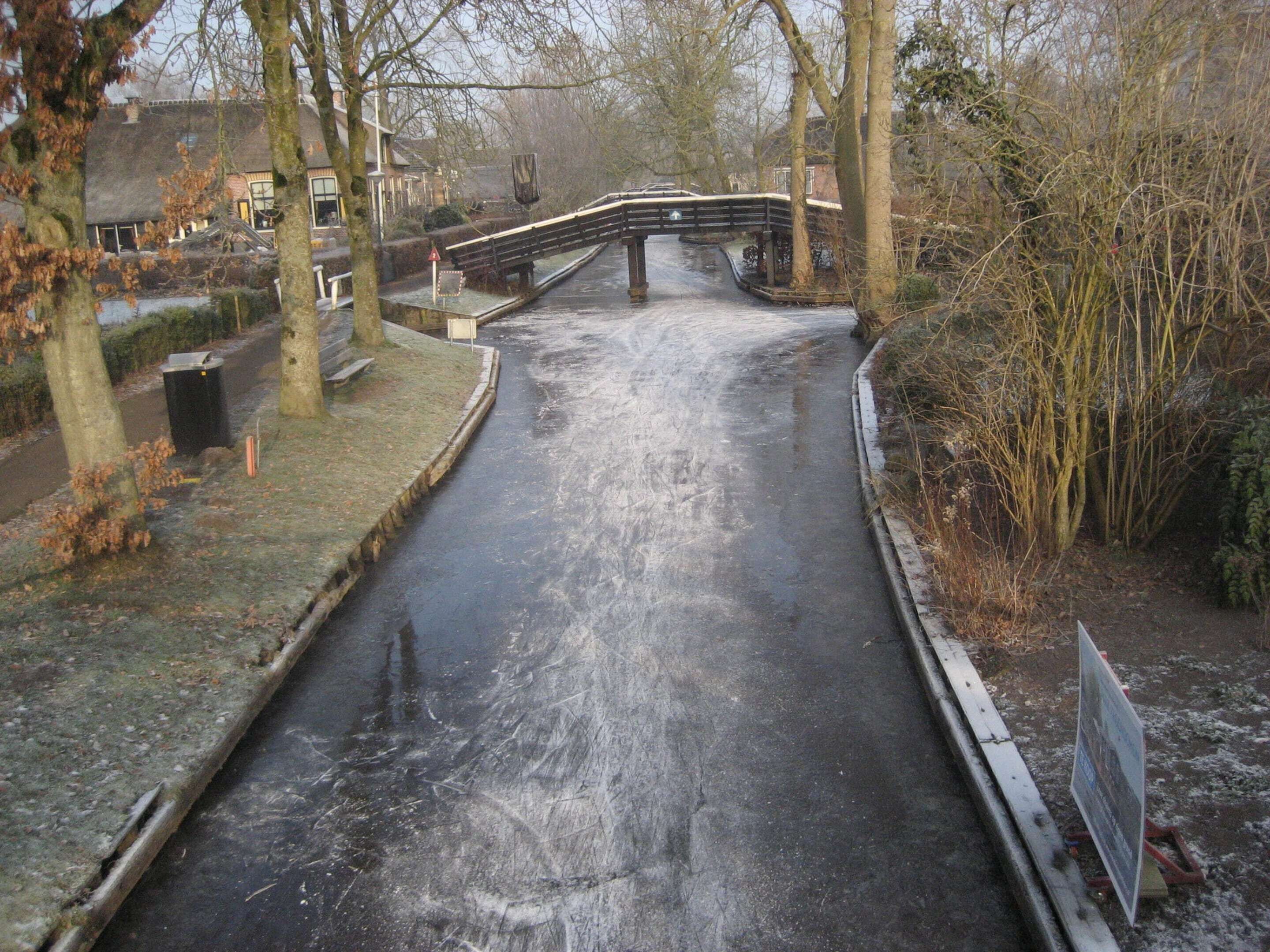 A narrow, icy canal with a bridge and trees on either side.