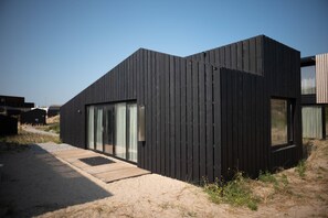 A modern black building with a wooden deck, large windows, and a sandy surrounding. - Modern Lodge With Dishwasher, Near the Beach (Zandvoort)